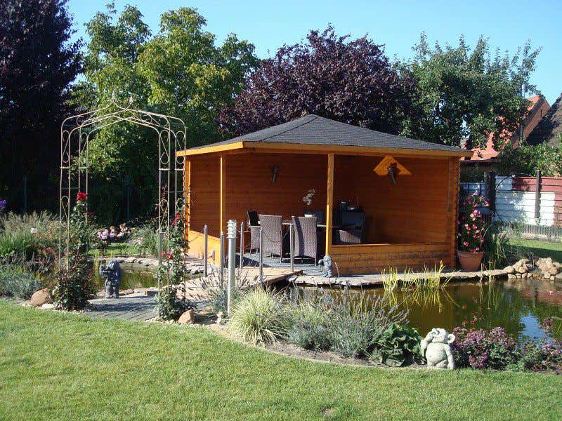 Mit einem offenen Gartenhaus sind Sie zwar draußen, gleichzeitig aber vor Wind und Wetter geschützt. Die 5-Eck Gartenlaube Maik ist charmant am Gartenteich platziert.